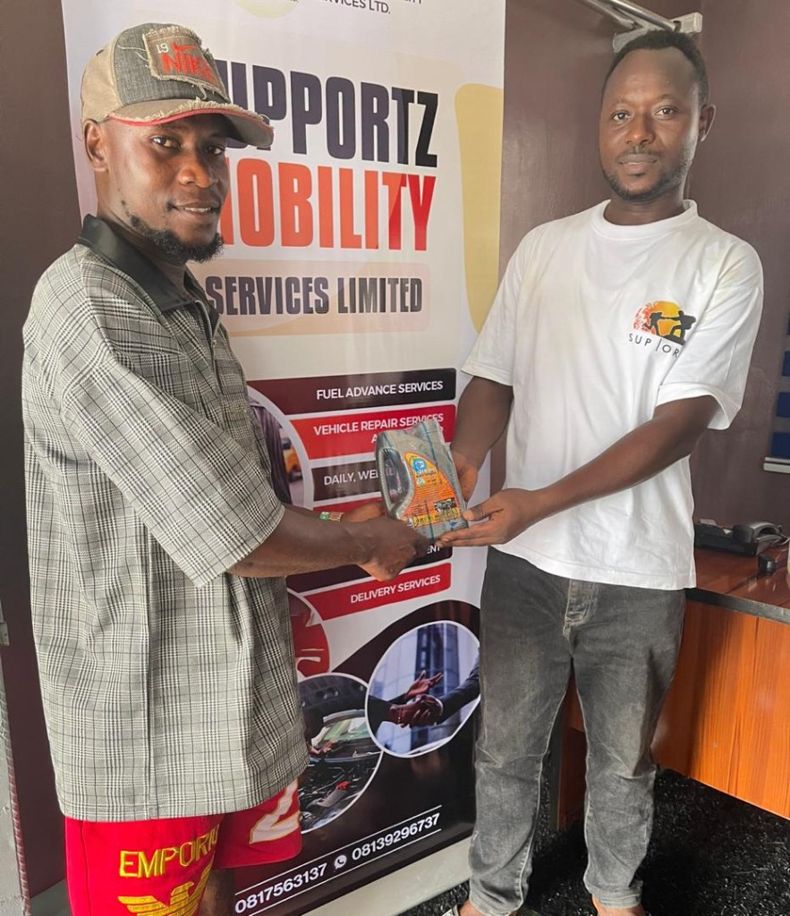 A Supportz staff member in a branded shirt handing motor oil to a driver in front of a Supportz Mobility Services banner